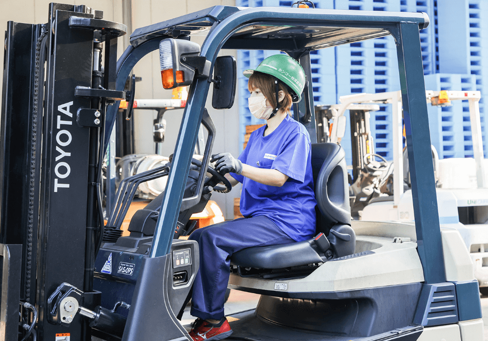 ヘルメットをかぶる女性スタッフがフォークリフトで荷物を運ぶ